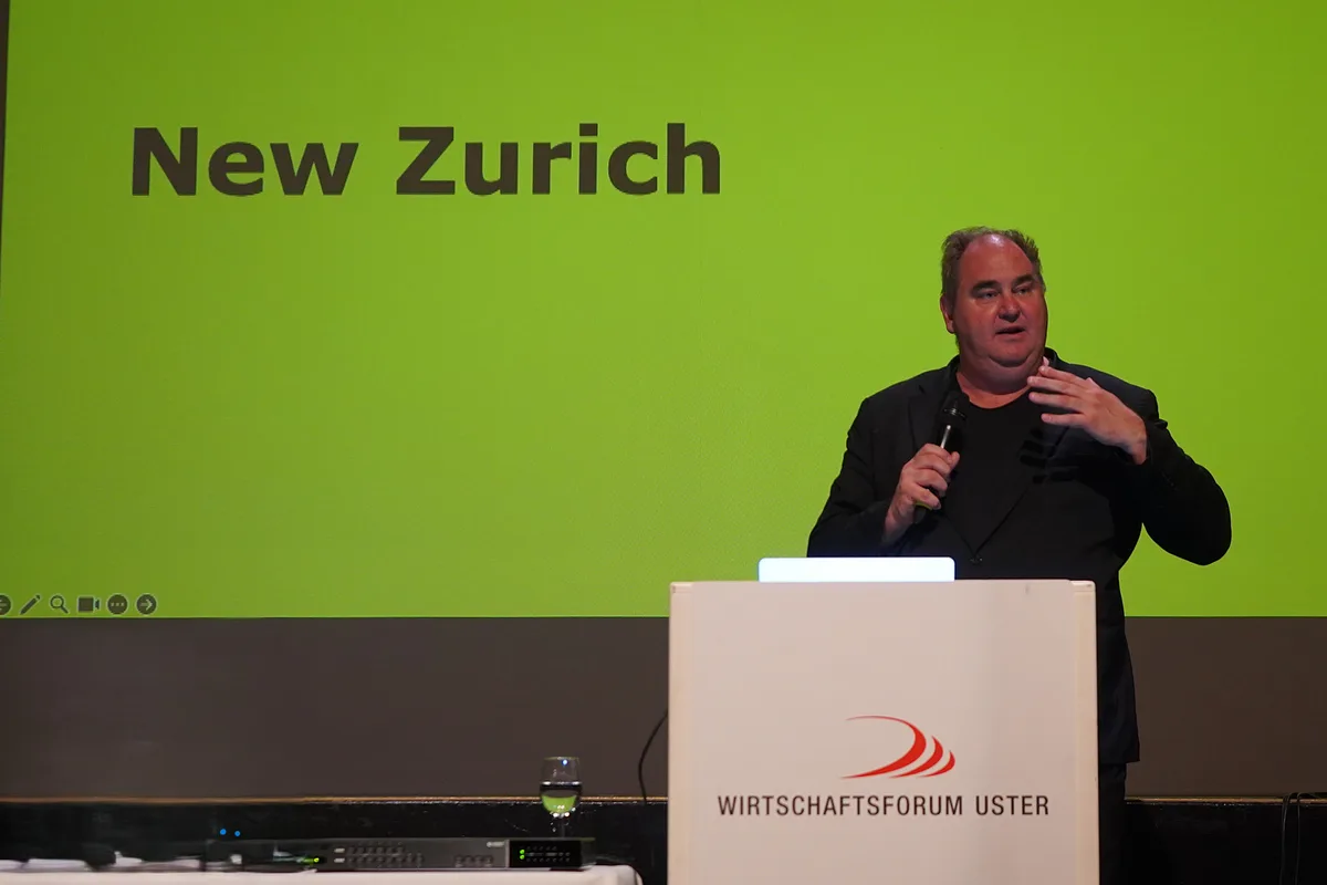 Ein Mann im dunklen Anzug steht auf einer Bühne und referiert. Im Hintergrund ist der Schriftzug «New Zurich» zu sehen.